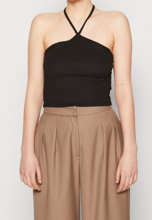 Osoba nosząca czarny bezrękawnik typu halter w stylu crop top w połączeniu z beżowymi, luźnymi spodniami z wysokim stanem na jednolitym tle.