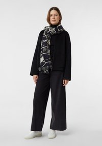 Kobieta w czarnych szerokich spodniach, czarnej kurtce na czarnym topie, wzorzystym czarno-białym szaliku i białych trampkach, stojąca na białym tle.