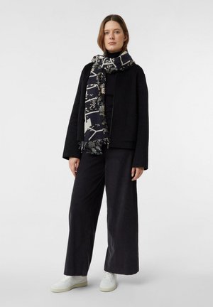 Kobieta w czarnych szerokich spodniach, czarnej kurtce na czarnym topie, wzorzystym czarno-białym szaliku i białych trampkach, stojąca na białym tle.