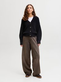 Woman with curly hair wearing a black buttoned cardigan over white shirt, loose brown pants, and dark brown shoes standing on white background.