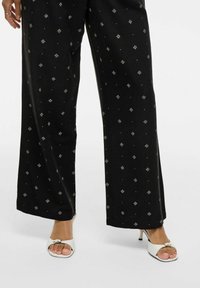 Pantaloni neri a gamba larga con motivi a diamante argentati, realizzati in tessuto morbido. Abbinati a tacchi bianchi open-toe con dettaglio a fibbia sulla parte anteriore.