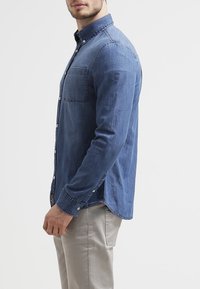 Camisa de mezclilla en azul oscuro, con cuello abotonado, bolsillo frontal y mangas largas. Textura suave con una silueta ajustada.