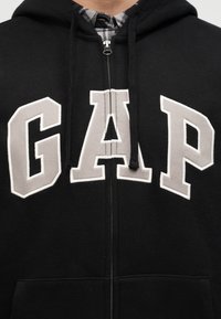 Sweat à capuche noir zippé avec de grandes lettres beiges "GAP" sur la poitrine et des cordons noirs, porté par-dessus un col de chemise à carreaux.