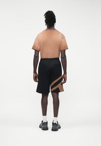 Hombre con rastas de espaldas que lleva una camiseta marrón deslavada, pantalones cortos negros con rayas diagonales marrones, calcetines blancos y zapatillas negras.