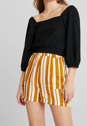 Femme portant un haut noir épaules dénudées avec des manches trois-quarts et un short taille haute orange et blanc à rayures verticales.