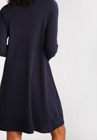 Abito in maglia di colore blu navy con maniche lunghe e design svasato. Tessuto liscio, lunghezza al ginocchio, con scollo rotondo e senza elementi hardware visibili.