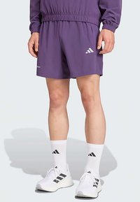 Fialové športové šortky s elastickým pásom, vybavené reflexnými pruhmi a logom Adidas. Doplnkované bielymi ponožkami a topánkami.