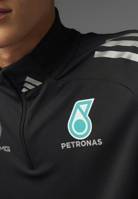 Sort lynlåsjakke med høj krave, der har sølvstribede detaljer på skulderen og et turkist "PETRONAS" logo på brystet.