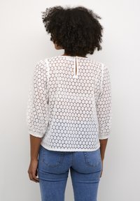 Witte kanten top met een honingraatpatroon, 3/4 mouwen en een keyhole-opening aan de achterkant, gecombineerd met blauwe jeans.