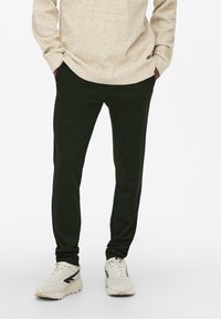 Donkergroene taps toelopende broek met een gladde textuur, gecombineerd met een lichtbeige shirt met lange mouwen en witte sneakers met zwarte accenten.