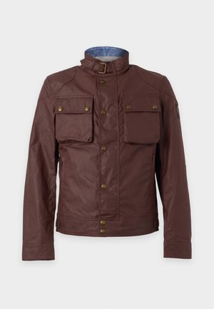 Homme portant une veste marron avec des poches à boutons, une chemise noire, un jean bleu et des chaussures en cuir noir, debout devant un fond uni.