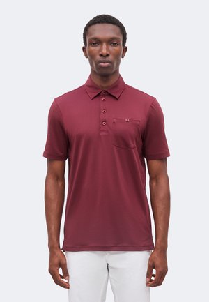 Homme portant un polo bordeaux à manches courtes avec une poche poitrine boutonnée et un pantalon blanc, debout devant un fond clair uni.