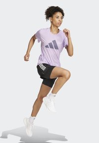 Violet lyhythihainen urheilupaita, jossa on harmaa Adidas-logo, yhdistettynä mustiin shortseihin, joissa on kolme valkoista raitaa. Valkoiset urheilukengät ja sukat.