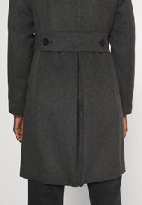 Szary wełniany płaszcz o prostym kroju, z rozcięciem z tyłu i dwoma czarnymi guzikami. Posiada długie rękawy i teksturowane wykończenie.