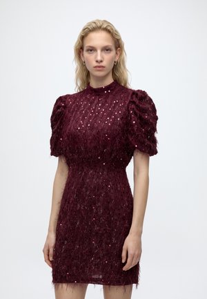 Blonde woman wearing a short burgundy dress with puffed sleeves and sequin details, standing against a plain white background.