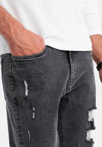 Hombre que lleva puesto unos vaqueros negros desgastados, con la mano en el bolsillo y una camisa blanca de manga larga, mostrando una muñeca con una pulsera de cuentas negras.