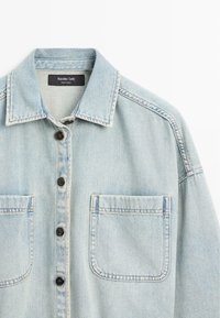 Lichtblauwe spijkerjasje met knopen aan de voorkant, twee borstzakken en een kraag, gelabeld Massimo Dutti, gemaakt in Turkije.