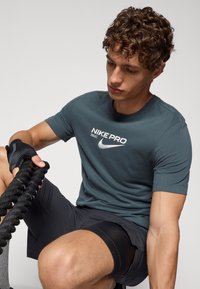 Nike Pro sportinis marškinėliai tamsiai žalia spalva, su balta logotipu ir tekstu. Derinami su juodais šortais. Matomi laikikliai ir storas virvė.