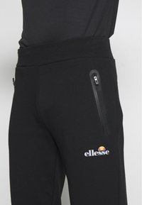 Black joggers made of cotton blend, featuring a front zip pocket, ribbed waistband, and a white logo on the left thigh. Smooth texture.