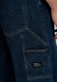 Pantalones de mezclilla azul oscuro con costuras reforzadas, un bolsillo de parche y una etiqueta. La tela tiene un acabado texturizado y un diseño de pierna recta.