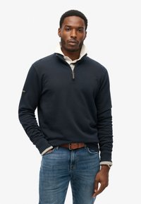 Sudadera navy con cuello de cremallera, con textura acanalada. Superpuesta a una camisa blanca con cuello. Combinada con jeans azules y un cinturón marrón.