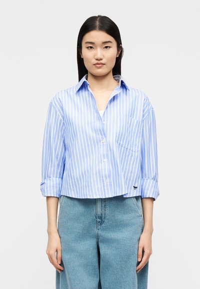 Femme aux cheveux noirs raides portant une chemise à boutons à rayures verticales bleues et blanches avec les manches retroussées et un jean bleu clair ample.