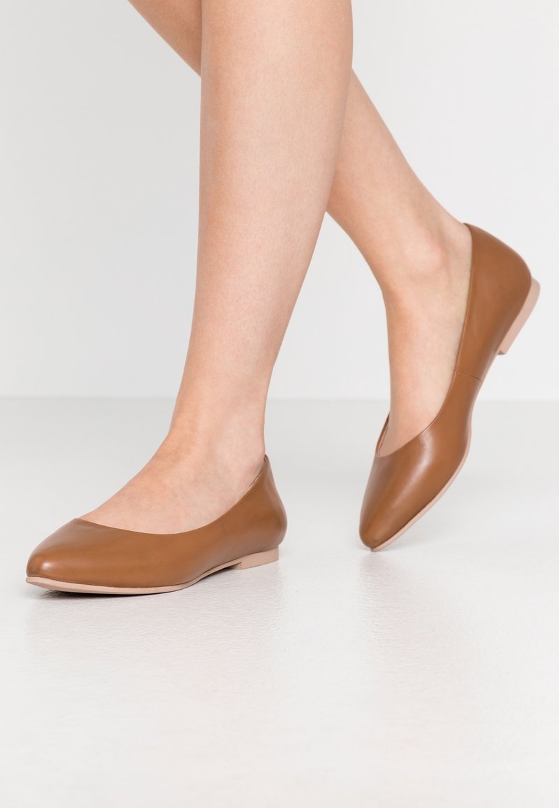 Femme portant des ballerines en cuir marron lisse, debout sur un sol clair avec un pied légèrement levé.