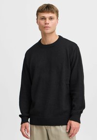 Suéter negro de punto con cuello redondo, mangas largas y puños acanalados, que presenta una tela texturizada sin patrones. Fondo neutro.