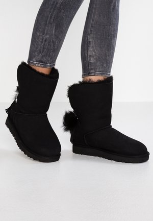 Nogi w wyblakłych czarnych obcisłych dżinsach, noszące czarne zimowe buty za kostkę z futrzanym wykończeniem i ozdobnymi pomponami.