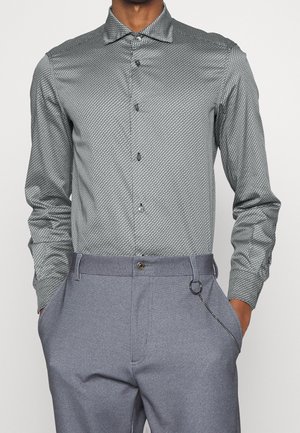 Homme portant une chemise grise ajustée à manches longues à motifs, boutonnée et rentrée dans un pantalon gris clair, les mains dans les poches.