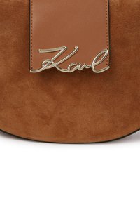 Brun ruskind håndtaske med en iøjnefaldende tan læderklap og et metalisk sølvfarvet signatur "Karl" logo. Glat tekstur med rund form.