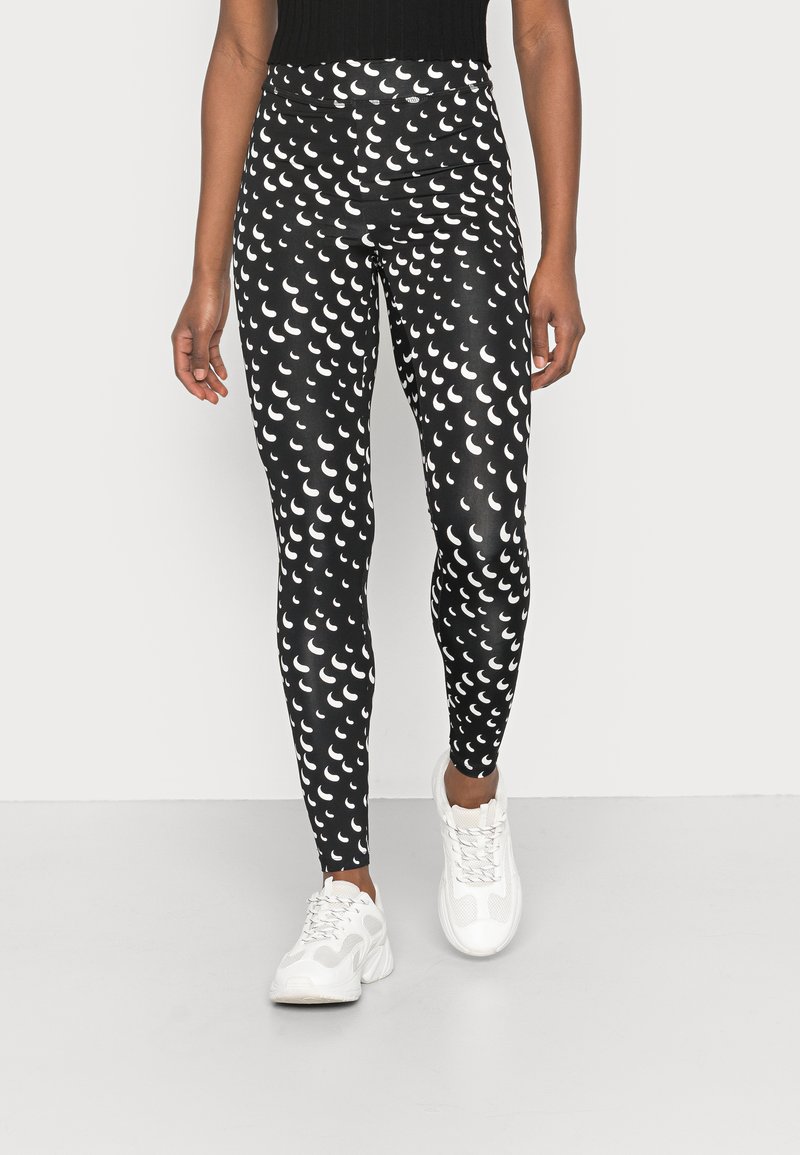 Leggings pretos em estilo ajustado, com um padrão de formas de crescente brancas, feitos de material elástico, combinados com ténis brancos.