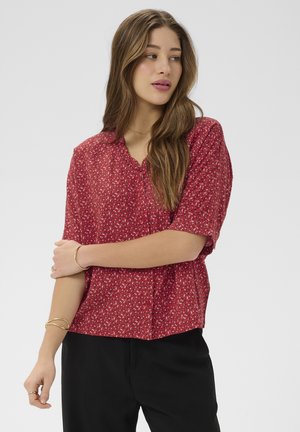 Mujer con cabello largo y castaño, vistiendo una blusa roja de manga corta con pequeños patrones blancos y pantalones negros, mirando hacia su izquierda.