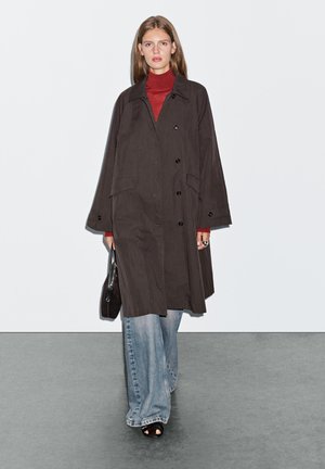 Manteau noir long avec un design à boutons, poches avant et manches larges, associé à un col roulé rouge et un jean bleu à jambes larges.
