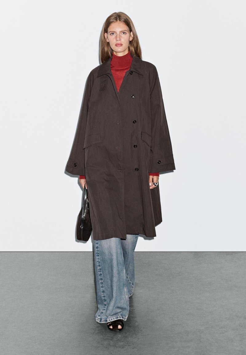 Schwarzer, langer Mantel mit einem Knopf-Design, Fronttaschen und weiten Ärmeln, kombiniert mit einem roten Rollkragenpullover und blauen weiten Jeans.