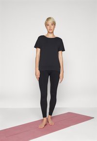 Maglietta nera a maniche corte con colletto rotondo, abbinata a leggings neri a coste, su un tappetino da yoga rosa. La modella è in piedi a piedi nudi, con le braccia rilassate ai lati.