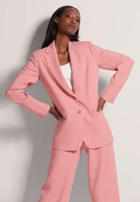 Roze pak bestaande uit een op maat gemaakte blazer met een sluiting met twee knopen, notch revers en wijde broeken, gemaakt van een gladde stof.