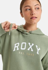Green cotton hoodie with a front graphic. Features white "ROXY" text and "EST. 1990" in gold. Adjustable drawstrings with metal tips.