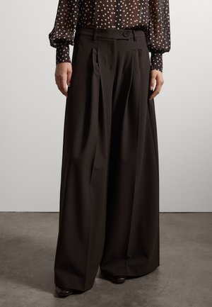 Persoon die zwarte wijde broek en een zwarte blouse met witte stippen en lange mouwen draagt, staande op een grijze vloer.