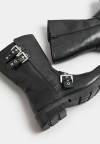 Botas de tobillo de cuero negro con dos hebillas plateadas, una superficie texturizada y una suela gruesa con dibujo. Cierre lateral con cremallera para un uso fácil.