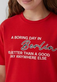 Rode katoenen t-shirt met de tekst "EEN SAAIETAG IN Berlijn IS BETER DAN EEN GOEDE DAG GENDAAR ANDERS" in contrasterende zwart-witte letters.