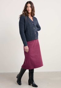 Marineblauwe kabelgebreide cardigan met houten knopen, gecombineerd met een bordeauxrode rok en zwarte kniehoge haklaarzen, op een neutrale achtergrond.