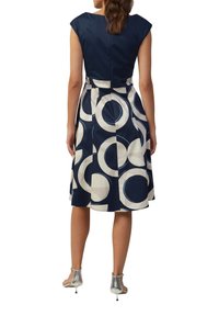 Kleid in Marineblau und Creme mit Kapuzensleeves, kreisförmigen Mustern und einem taillierten Oberteil sowie einem schwingenden Rock. Silberschicke High Heels runden den Look ab.