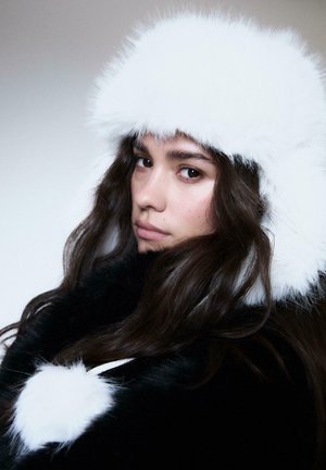 Ung kvinde med langt mørkt hår, iført en hvid fluffy hat og en matchende sort frakke med hvide pom-pom detaljer, der ser lidt til siden.