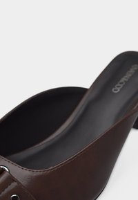 Καφέ δερμάτινο μοκασίνι με χαμηλό τακούνι και μαύρη επένδυση με μαξιλαράκι, που διαθέτει ασημένια μεταλλική αγκράφα στο πάνω λουράκι.