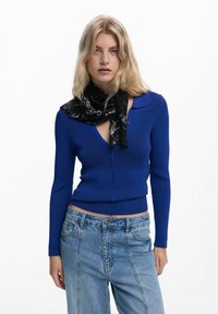 Sininen röyhelö cardigan kauluksella, syvällä V-pääntiellä ja nappisuljin, yhdistetty mustan kuviollisen huivin ja farkkuhousujen kanssa.