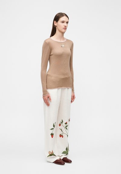 Kvinde stående iført en gennemsigtig beige langærmet top og hvide bukser med vide ben med broderi af jordbær og snegl, kombineret med brune sko.