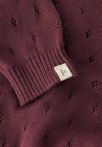 Bordo spalvos megztinis su tekstūruotu raštu, su ribinėmis rankogaliais ir mažyte etikete siūta prie apačios. Pagamintas iš minkšto vilnos mišinio.