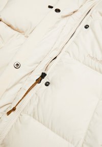 Beige pufferjack met een doorstikt patroon, voorzien van een ritssluiting met gouden accenten en zwarte knopen. Geborduurd logo in de stof.