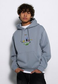 Jasnoszara bluza z kapturem i przednią kieszenią, z kolorowym haftowanym wzorem z napisem "LOKALNI BOHATEROWIE GRAJĄ" oraz różnymi ikonami.